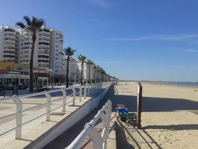 a beach with buildings and palm trees and a white fence at Apartamento Las Olas de Valdelagrana in El Puerto de Santa María
