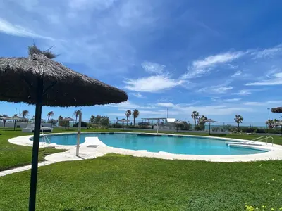 a large swimming pool with an umbrella and grass at Moderno Apartamento Primera Línea Playa de La Barrosa by ChiclanaDreams in Chiclana de la Frontera