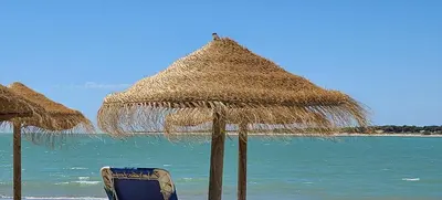 two straw umbrellas and a chair on a beach at Apartamento JARDINES DE BAJO GUIA in Sanlúcar de Barrameda