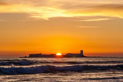 a sunset over the ocean with a lighthouse in the distance at Pipoca's Beach in Novo Sancti Petri