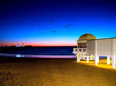 a flock of birds flying over a building on the beach at Playa La Caleta in Cádiz