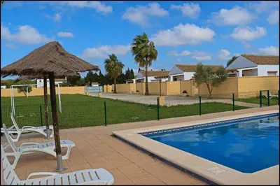 a pool with a straw umbrella and two chairs and a table at Bungalow en complejo II in Conil de la Frontera
