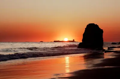 a sunset on the beach with a large rock in the water at Apartamento Plaza Castañuelas in Jerez de la Frontera