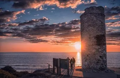 two people standing next to a stone tower near the ocean at El Ancla in Chiclana de la Frontera